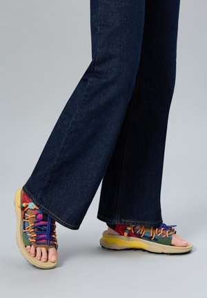 Piedi con sandali colorati aperti con lacci, abbinati a jeans larghi blu scuro, su uno sfondo grigio neutro.