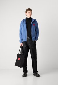 Blå hettejakke med rød logo, svart høyhalset genser, svarte vide jeans, svart totebag med rød etikett, svarte sko, minimalistisk bakgrunn.