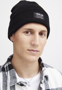 Musta ribbed-pipo, jossa on merkki "!SOLID". Henkilöllä on vaalea iho, mustavalkoinen ruudullinen takki ja hopeakorut.