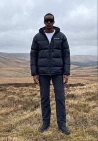 Férfi fekete puffert dzsekit, sötét nadrágot és napszemüveget viselve, gyepes dombon áll, háttérben hullámzó dombokkal és felhős éggel.