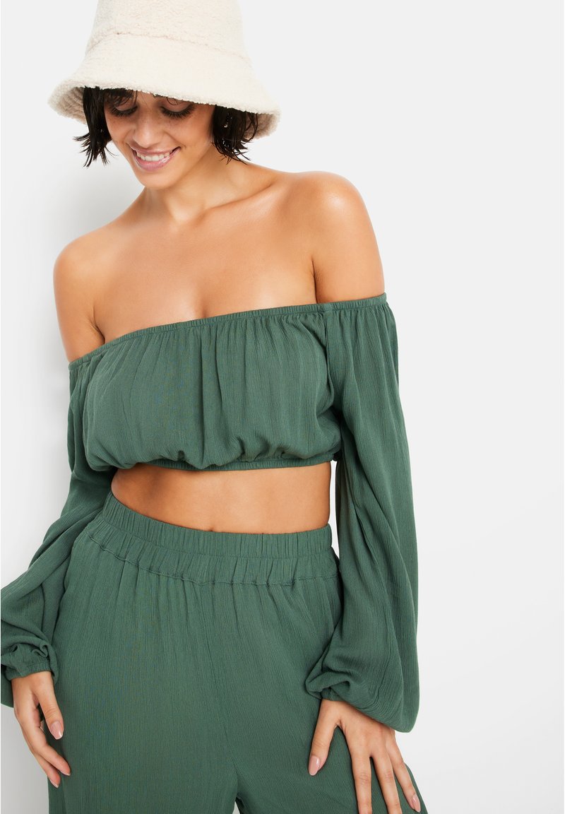 Πράσινη off-the-shoulder cropped μπλούζα με ελαστική μέση και μακριά, ρέουσα μανίκια, σε συνδυασμό με άνετους παντελόνες σε χαλαρή γραμμή. Μαλακή υφή.