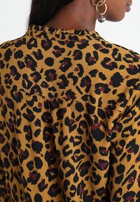 Blusa in stampa leopardata gialla senape, con macchie nere e borgogna, vestibilità ampia, tessuto testurizzato e dettaglio di scollatura arricciata.
