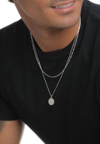 Due collane d'argento sovrapposte sopra una maglietta nera. Una ha un ciondolo rotondo con superficie testurizzata, l'altra presenta un delicato disegno a catena.