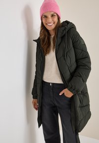 Grön pufferjacka med dragkedja och huva, matchad med en beige tröja och mörkgrå byxor. Modellen bär en ljusrosa stickad mössa.