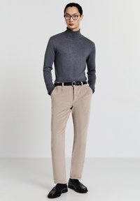 Maglione grigio a collo alto, pantaloni in corduroy beige, cintura nera e scarpe nere. Il modello si trova con le mani in tasca contro uno sfondo neutro.