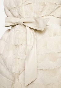 Płaszcz z sztucznego futra w kolorze kremowym z teksturowanym, warstwowym wzorem; posiada gładki, szeroki pasek związany w kokardę w talii, co podkreśla sylwetkę.