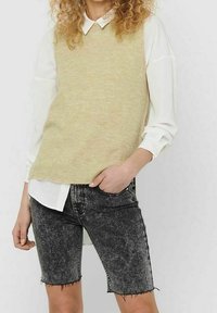 Gilet a maglia giallo chiaro sopra una camicia bianca con colletto, abbinato a pantaloncini in denim neri con orli sfilacciati e una texture sbiadita.