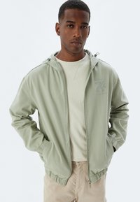 Lys grøn nylonjakke med hætte, elastiske manchetter og talje. Har et lille logo på brystet og bæres over en beige crew-neck skjorte.
