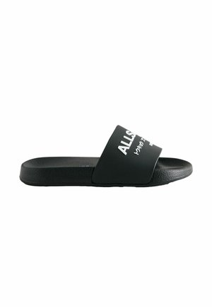 Svart gummislip-sandal med en bred rem som har vit text, platt sula, texturerad fotdel och minimalistisk design.