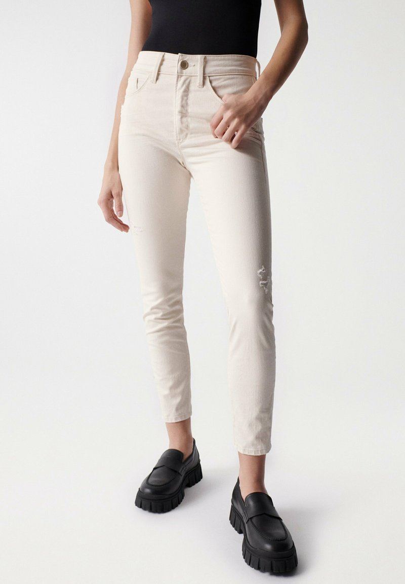 Jeans skinny de color beige claro con cintura alta, que presentan detalles desgastados y diseño de cinco bolsillos. Combinados con loafers negros gruesos.