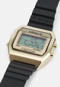 Digitális óra arany fém házzal, fekete gumi szíjjal és LCD kijelzővel, amelyen az idő és a nap látható. Két gomb található az oldalán.