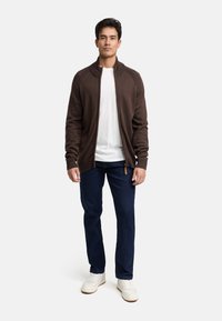 Brązowy kardigan na zamek błyskawiczny z prążkowanym kołnierzem, noszony na białej koszulce, zestawiony z ciemnoniebieskimi dżinsami i białymi sneakersami. Luźny krój.