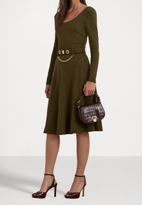 Olivově zelené šaty s dlouhým rukávem a přiléhavým korzetem, rozšířenou sukní, doplněné zlatým řetízkovým páskem. Hnědá kabelka s krokodýlím vzorem a vysoké podpatky.