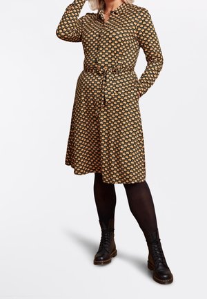 Femme portant une robe à manches longues à motifs avec une ceinture à la taille, des collants noirs et des bottes noires lacées, debout une main sur la tête.