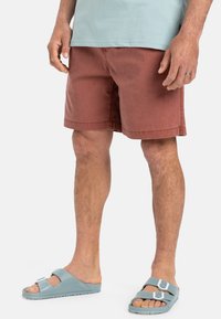 Férfi, rozsda színű, pamut shorts, egyenes szabású, oldalsó zsebekkel. Világoskék, dupla pántos szandállal párosítva.
