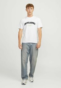 Camiseta blanca de algodón con el logo negro "JACK&JONES", combinada con jeans claros de corte holgado y zapatillas deportivas plateadas.