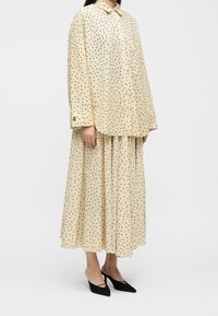 Femme portant un chemisier ample crème et une jupe plissée assortie à pois noirs, associée à des chaussures slingback noires à bout pointu.