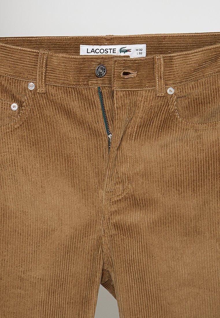 Pantalon en velours côtelé marron, avec une texture à côtes verticales, fermeture éclair, boutons en argent et logo Lacoste sur la ceinture.