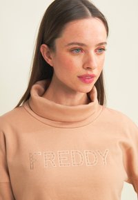 Giovane donna con lunghi capelli castani che indossa una felpa beige con collo alto con il testo "FREDDY" sulla parte anteriore, che guarda leggermente verso destra.