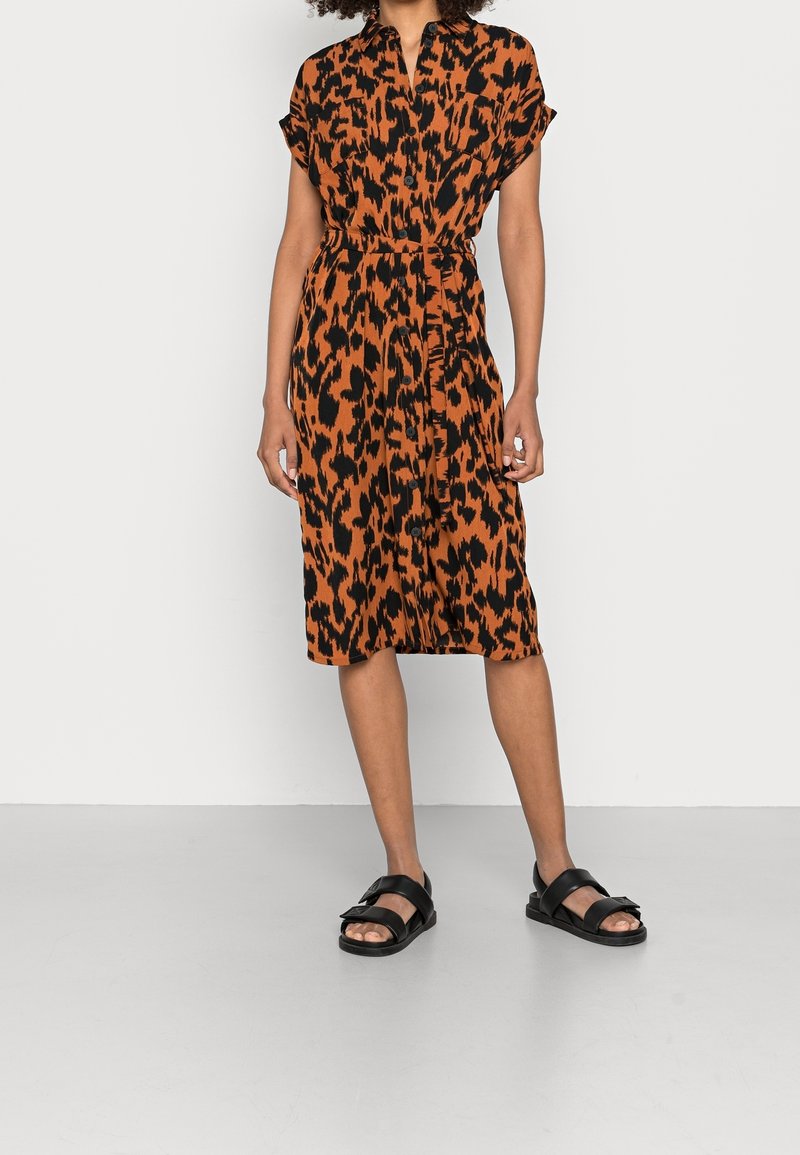 Femme portant une robe orange longueur genou avec des taches abstraites noires, manches courtes, boutons sur le devant et sandales noires à brides.