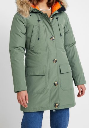 Femme portant un manteau d'hiver vert à capuche avec bordure en fourrure, doublure orange, grands boutons, poches avant, et jean bleu.