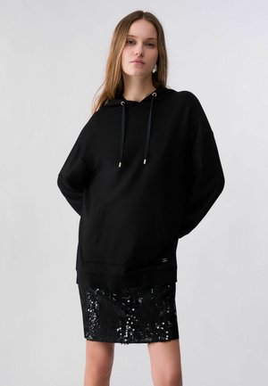 Junge Frau trägt einen schwarzen übergroßen Hoodie und einen kniehohen schwarzen Paillettenrock und steht vor einem einfachen Hintergrund.