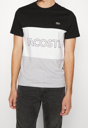 Un homme portant un t-shirt Lacoste en noir, blanc et gris avec un design en blocs de couleurs, affichant un grand texte de marque et un petit logo de crocodile sur la poitrine.