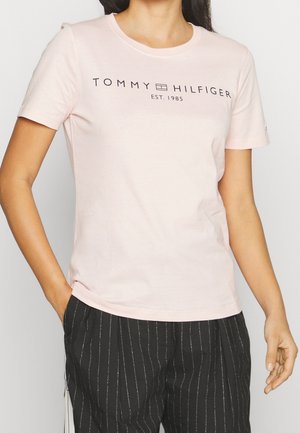 Kobieta ubrana w jasnoróżową koszulkę Tommy Hilfiger z logo i czarne spodnie w cienkie paski, z jedną ręką w kieszeni, stojąca na jasnym tle.