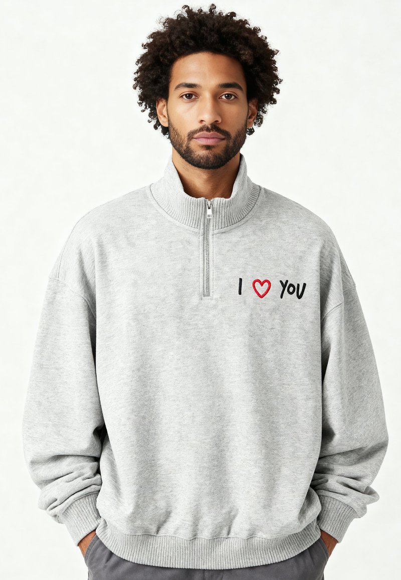 Sweat-shirt gris à fermeture éclair avec col montant, présentant le texte brodé "I ❤️ YOU" en noir et rouge sur la poitrine gauche, avec des poignets côtelés.