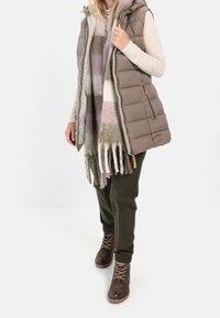 Gilet marrone imbottito con cappuccio, maglione leggero, sciarpa a quadri con frange, pantaloni verdi a sigaretta e stivali marroni stringati con finitura testurizzata.