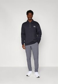 Zwarte sweatvest met ritssluiting en wit logo, gecombineerd met grijze broek en witte sneakers. Model staat tegen een lichte achtergrond.