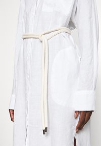 Una persona indossa una lunga camicia di lino bianco, stretta in vita con una cintura di corda di colore chiaro, mostrando tasche visibili e dettagli della camicia.