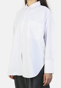 White long-sleeve button-up shirt featuring a collar, a front pocket, and a curved hem, made of smooth fabric with a slight sheen.