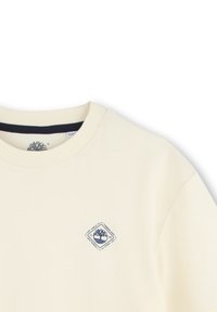 Crème-kleurige sweatshirt met een ronde hals, korte mouwen en een marineblauw diamanten logo op de linker borst. Zachte, gladde textuur.