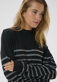 Blonde woman wearing black sweater with horizontal silver stripes, arms crossed, looking to the side with neutral expression on a light background.