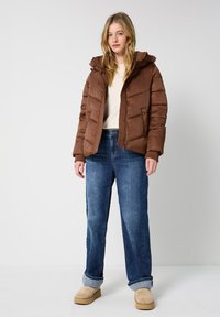 Giacca a vento marrone con cappuccio, design trapuntato e polsini a coste, indossata sopra un maglione color crema, abbinata a jeans a gamba larga blu chiaro e scarpe beige.