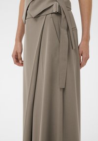 Taupe slå-om-nederdel med sidelukning og integreret bælte, der viser folder og sidelomme, båret af en person med afslappede arme.