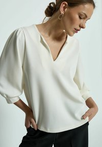 Ivoren V-halsblouse met puffmouwen van zachte stof. Bevat opgerolde manchetten en een relaxte pasvorm, gecombineerd met zwarte broeken.