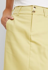 Mano descansando en el bolsillo de unos pantalones de tiro alto de color amarillo claro con dobles botones dorados y lazos para el cinturón, llevados con una camiseta blanca.