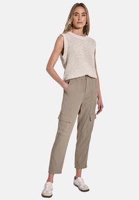 Beige stickad ärmlös topp som kombineras med khaki cargo-byxor med sidofickor, bärs tillsammans med beige och vita sneakers framför en enkel bakgrund.