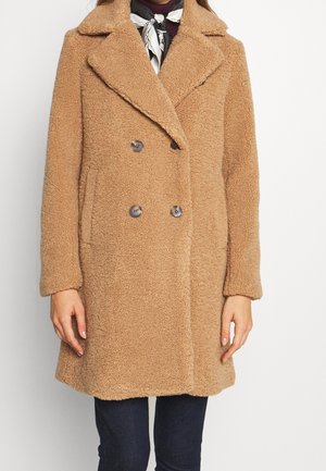 Femme portant un manteau teddy beige mi-long avec de grands revers, des boutons croisés et des poches latérales, sur un pantalon foncé et un foulard en soie autour du cou.
