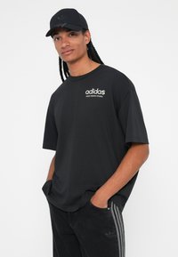 Črna bombažna majica s kratkimi rokavi in okroglim izrezom, s belim logotipom "adidas". Ujemajoče temne kavbojke in kapica.
