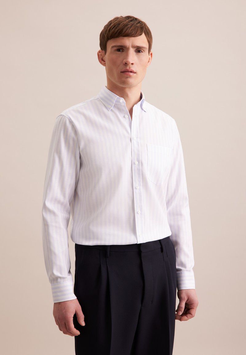 Witte blouse met lichtblauwe verticale strepen, lange mouwen en een linkerschouderzak, gecombineerd met donkere broeken. Vlotte textuur.