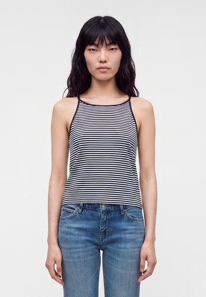 Femme aux longs cheveux noirs portant un débardeur rayé noir et blanc et un jean bleu, debout devant un fond clair uni.