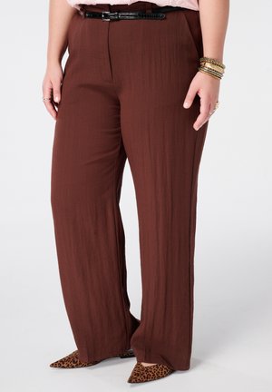 Personne portant un pantalon marron ample avec ceinture noire, chaussures pointues imprimé léopard et bracelets dorés au poignet droit.
