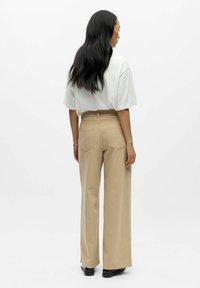 Bēšas platām kājām bikses ar augstu vidukli, kurām ir divas aizmugures kabatas un gluda tekstūra. Kombinētas ar vaļēju baltu kreklu.