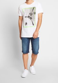 Hvid bomulds T-shirt med grafikprint og grøn kant, parret med knælange denim shorts og hvide sneakers.