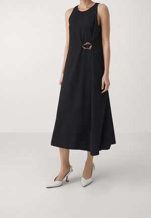 Kobieta ubrana w czarną midi sukienkę bez rękawów z ozdobą w postaci złotego łańcucha w talii oraz białe szpilki na obcasie z odkrytą piętą i ostrym noskiem.