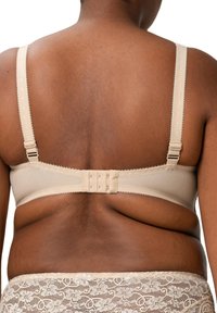 Beige rintaliivi, jossa on säädettävät hihnat ja koukku-silmukkakiinnitys. Liukas materiaali, aaltoileva trimmi hihnojen reunoilla ja teksturoitu pitsivyöt. 