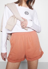Orange bomullsshorts med uppvikta fållar och sidofickor, kombinerade med en vit långärmad skjorta med en svart logotyp. Beige tröja draperad.
